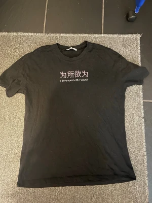 Svart t-shirt med tryck - Svart t-shirt med korta ärmar och rund hals. På bröstet finns ett tryck med kinesiska tecken och texten 'I DO WHATEVER I WANT' i vitt och rosa. Perfekt för dig som gillar att sticka ut med coola statement-plagg.