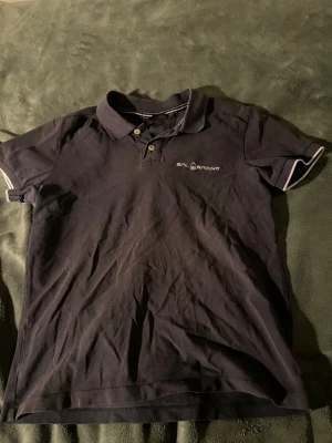 Svart piké från Sail Racing - Säljer en svart pikétröja från Sail Racing med klassisk krage och två knappar framtill. Diskret logga broderad på bröstet och vita ränder på ärmsluten. Tröjan är gjord i mjuk bomull och har korta ärmar, perfekt för en chill och sportig look.