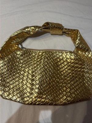 Guldig axelväska med krokodilmönster - Säljer en snygg axelväska i guld med krokodilinspirerat mönster. Väskan har en bred axelrem med knutdetalj och stängs med dragkedja. Perfekt för dig som vill sticka ut med en unik accessoar. Materialet är glansigt och ger en lyxig känsla.