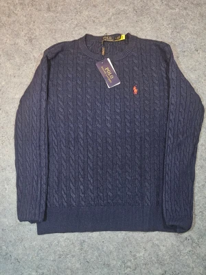 Marinblå stickad tröja Polo Ralph Lauren - Snygg marinblå stickad tröja från Polo Ralph Lauren i storlek S. Tröjan har ett klassiskt kabelstickat mönster och en röd broderad logga på bröstet. Rund halsringning och långa ärmar ger en stilren look. Perfekt för dig som gillar tidlös design och premiumkänsla.
