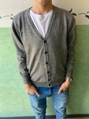 Grå kofta från Gianni Lupo, M - Snygg grå kofta från Gianni Lupo i storlek M. Koftan har v-ringad hals, långa ärmar och svarta knappar framtill. Materialet är mjukt och tunt, perfekt för lager-på-lager. Passformen är normal och den neutrala färgen gör den enkel att matcha med jeans eller t-shirt.