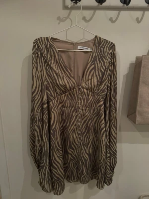 Zebramönstrad klänning från Adoore - Säljer en snygg zebramönstrad klänning från Adoore i beige och brunt. Klänningen har lång ärm, v-ringad hals och knappar framtill. Materialet känns lätt och luftigt, perfekt för dig som gillar djurmönster och vill sticka ut.