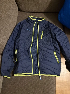 Mörkblå pufferjacka från Bergans - Säljer en mörkblå pufferjacka från Bergans med coola gula detaljer och dragkedjor. Jackan har quiltad design, hög krage och tre fickor med dragkedja. Perfekt för dig som gillar outdoor och vill ha en snygg jacka med sportig vibe.