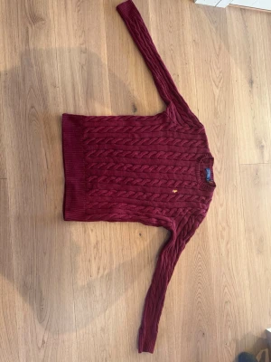 Vinröd stickad tröja Polo Ralph Lauren - Säljer en vinröd stickad tröja från Polo Ralph Lauren med klassiskt kabelstickat mönster och gul broderad logga på bröstet. Tröjan har rund hals och långa ärmar. Perfekt för dig som gillar stilrena och tidlösa plagg.