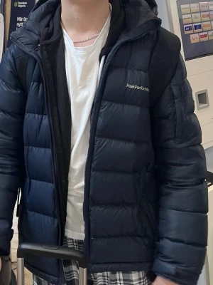Mörkblå Peak Performance dunjacka - Säljer en mörkblå dunjacka från Peak Performance med huva och dragkedja framtill. Första bilden är mest lik verklig färg.  Jackan har quiltad design och är tillverkad i syntetmaterial med dunfyllning. Perfekt för kalla dagar och har en clean look med logga på bröstet.