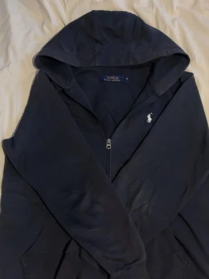 Blå Ralph Lauren Zip Hoodie - Blå Ralph Lauren Zip Hoodie som säljs pga att den inte används tillräckligt. Tröjan är köpt här på plick och är i bra skick! Det är bra kvalité och den är väldigt snygg och skön😁 tröjan är använd 10-15 gånger. Hör gärna av er i DM om ni har några funderingar!