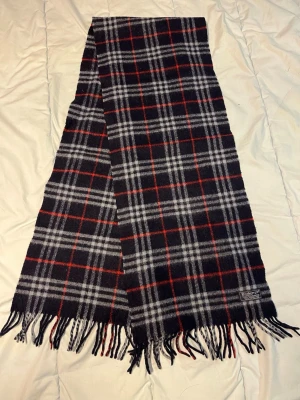 Rutig halsduk  från Burberrys - Säljer en klassisk rutig halsduk från Burberrys i mörkblå, grå och röd färg. Halsduken är tillverkad i 100% lammull och har fransar längs nederkanten. Perfekt för dig som gillar tidlös design och vill ha något som sticker ut lite extra.