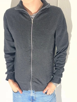 Mörkgrå kofta från Jack & Jones - Säljer en mörkgrå stickad kofta från Jack & Jones Originals i storlek medium. Koftan har hel dragkedja framtill, ribbade muddar och krage. Materialet är mjukt och har en diskret struktur på överdelen. Perfekt för dig som gillar stilrena och bekväma plagg.