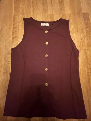 Vinröd ärmlös topp från Holly & Whyte - Snygg vinröd ärmlös topp från Holly & Whyte by Lindex i storlek XS. Toppen har rund hals och dekorativa guldknappar framtill. Materialet känns mjukt och har en lätt struktur. Perfekt att styla med jeans eller kjol för en cool look. Aldrig använd