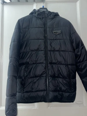 Svart pufferjacka Supply & Demand - Svart pufferjacka från Supply & Demand med huva och dragkedja framtill. Jackan har quiltad design, två sidofickor med dragkedja och en snygg logotyp på bröstet. Insidan är ljusgrå och materialet känns lätt och syntetiskt. Perfekt för kyliga dagar. Du kan vända jackan på andra sidan så får du en ”reflex” aktig färg. Den är alltså tvåsidig där du kan använda både sidorna beroende på vilken färg du känner för.