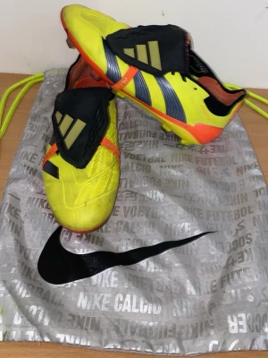 Adidas Predator gula fotbollsskor - Säljer ett par Adidas Predator fotbollsskor i storlek 40. Skorna är gula med svarta och orange detaljer, har täckt snörning och en cool Predator-logga under sulan. Ovandelen är i syntetmaterial och designen är riktigt sportig och modern. Perfekt för dig som vill dominera på planen. Den kommer också med benskydden i bilden, ett par blåa Mercurial benskydd med lila detaljer. Pris kan diskuteras. 