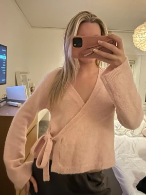 Ljusrosa omlottkofta med knytband - Supermysig ljusrosa omlottkofta med knytband i midjan. Koftan har lång ärm och är stickad i ett mjukt material som känns skönt mot huden. Perfekt att slänga på sig när det är lite kyligt ute.