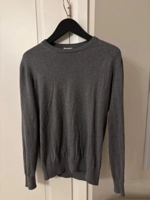 Grå stickad tröja J.Lindeberg L - Snygg grå stickad tröja från J.Lindeberg i storlek L men sitter som M. Tröjan har rund hals, ribbade muddar och mörkbruna förstärkningar på armbågarna. Perfekt för dig som gillar stilrena och klassiska plagg med en twist.