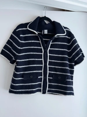 Randig kofta från Divided H&M, marinblå - Kortärmad kofta från Divided H&M i marinblått med vita horisontella ränder. Tröjan har bred krage, knäppning framtill med fyra knappar och två små fickor med knappdetaljer. Mjuk stickad kvalitet som känns chill och enkel att styla.