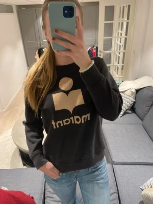 Svart sweatshirt från Isabel Marant - Cool svart sweatshirt från Isabel Marant Étoile med stort beige logotryck framtill. Tröjan har rund hals, långa ärmar och ribbade muddar. Perfekt till jeans för en avslappnad look.