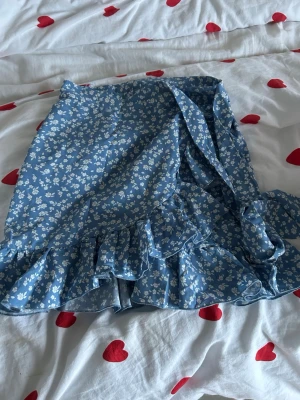 Blommig blå omlottkjol från SHEIN - Supersöt blå kortkjol från SHEIN i storlek XS med vitt blommönster. Kjolen har omlott-design och volangdetaljer längs nederkanten som ger en extra feminin touch. Perfekt för dig som gillar romantisk stil och vill ha något som sticker ut lite extra.