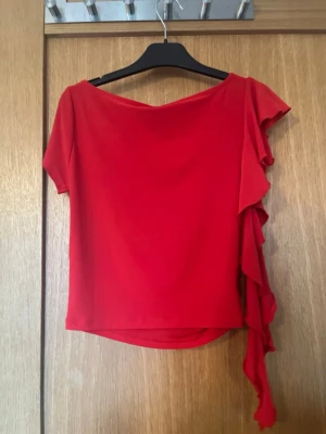 Röd festtopp med volang från Gina Tricot - Säljer en snygg röd topp från Gina Tricot i storlek S. Toppen har korta ärmar och en bred halsringning. Ena sidan är dekorerad med en lång volang som ger en cool asymmetrisk look. Materialet är mjukt och stretchigt, perfekt för att sticka ut. Används endast en gång