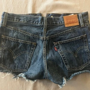 Levi's blå jeansshorts med fransar - Säljer ett par klassiska blå jeansshorts från Levi's med hög midja och råa, fransiga kanter. Shortsen har fem fickor, synliga knappar framtill och den ikoniska Levi's-lappen bak. Perfekta för en chill sommarlook.