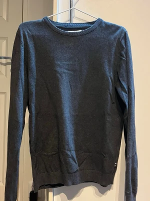 Mörkgrå långärmad tröja Jack & Jones - Säljer en stilren mörkgrå långärmad tröja från Jack & Jones i storlek XS. Tröjan har rund hals och ribbade muddar vid hals, ärmslut och nederkant. Materialet är mjukt och känns skönt mot huden, perfekt för kyligare dagar. Ett litet hål under armen