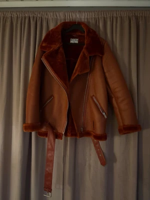 Brun teddyjacka från NA-KD - Säljer en brun teddyjacka från NA-KD med bred krage och mjukt foder. Jackan har dragkedjor på fickorna, ett bälte i midjan och detaljer i samma färg. Perfekt för dig som vill ha en cool och varm look under hösten och vintern. Använd endast ungefär 3 gånger max.