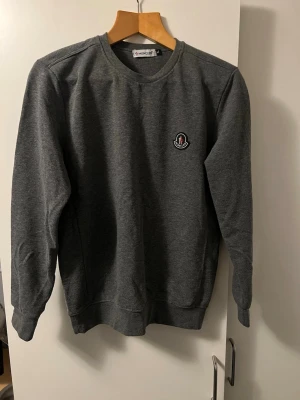 Grå sweatshirt från Moncler XL - Säljer en stilren grå sweatshirt från Moncler i storlek XL. Tröjan har rund hals, långärmade ärmar och ett snyggt Moncler-märke på bröstet. Perfekt för dig som gillar klassisk streetwear med exklusiv känsla.
