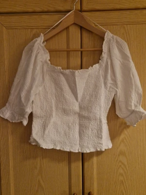 Vit blus med puffärmar från C - Säljer en vit blus från Cubus i storlek M. Blusen har puffiga trekvartsärmar, smockad struktur och söt volangkant vid halsringning och nederkant. Den är lätt och luftig, perfekt för varmare dagar. Passformen är normal och materialet känns som bomull. Endast provad, aldrig använd. Kolla gärna in mina andra annonser! 