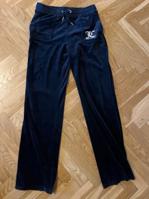 Juicy Couture mörkblå stl 15/16 år  - Mörkblå mjukisbyxor från Juicy Couture med logga på vänster ficka. Byxorna har snörning i midjan, raka ben och är gjorda i mjuk sammet. Perfekta för chill och häng, med skön loose passform och praktiska fickor fram och bak. Beninnerlängd 80 cm. Ej mkt använda! 