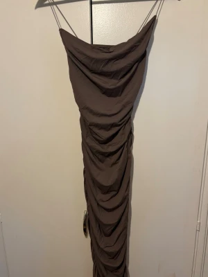Brun långklänning - Säljer en snygg brun långklänning med smala axelband. Modellen är figurnära och har en cool offshoulder-look upptill. 