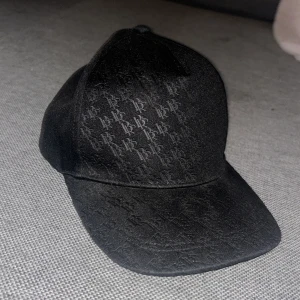 Svart keps med BB-mönster - Snygg svart keps med justerbar snapback och upprepade BB-loggor som mönster över hela kepsen. Materialet känns mjukt och har en matt finish. Perfekt för dig som vill ha en stilren accessoar med diskret branding.