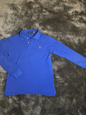 Blå långärmad piké från Polo Ralph Lauren - Säljer en blå långärmad pikétröja från Polo Ralph Lauren med orange broderad logga på bröstet. Tröjan har klassisk krage, två knappar vid halsen och är i en slim fit-modell. Perfekt för dig som gillar stilren och sportig look.