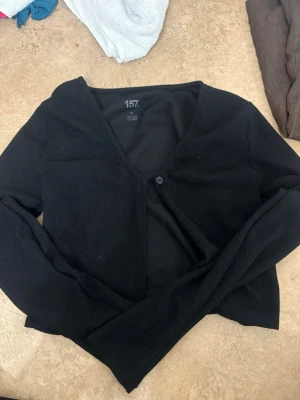 Svart ribbad kofta från 157 - Svart croppad kofta från 157 med ribbad struktur och v-ringad hals. Koftan har en knapp framtill och långa ärmar, perfekt att slänga över en t-shirt eller linne. Enkel och stilren design som funkar till många olika looks.