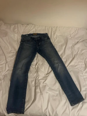 Blå jeans från Jack/Jones - Säljer ett par klassiska Jack/Jones jeans i blå tvätt med snygga slitningar. Modellen har fem fickor, normal passform och raka ben. Jeansen har en brun Jack/Jones lapp bak i midjan och är gjorda i slitstarkt denim-material. Perfekt för dig som gillar tidlös stil. Knappt använda och inget tecken på slitage. Storlek 29/30, nypris 1000 kr. Mitt pris 250 kr