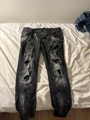 Svarta slitna jeans med färgstänk Dsquared 2 - Säljer ett par svarta jeans med rejält slitna hål och coola vita färgstänk över hela benen. Jeansen har en rak passform och detaljer som dragkedja vid fickan. Perfekta för dig som gillar en edgy och unik stil.