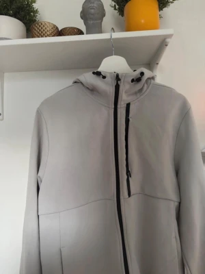 Ljusgrå vindjacka med huva - Säljer en ljusgrå vindjacka med huva och svarta dragkedjor. Jackan har justerbar snörning vid huvan och en clean, minimalistisk look. Perfekt för dig som gillar stilrena plagg med funktionella detaljer. Passar dig som vill ha en jacka med sportig vibe.