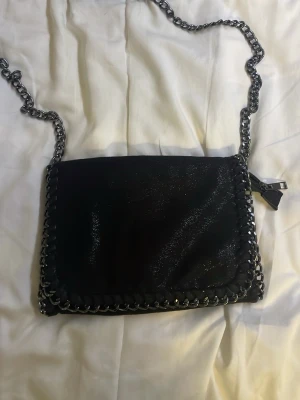Svart glittrig axelväska med kedja - Säljer en svart glittrig axelväska med grov silverfärgad kedja som axelrem och snygga kedjedetaljer runt kanten. Väskan har magnetlås och ett innerfack med dragkedja. Perfekt storlek för det viktigaste och ger en cool vibe till din outfit.