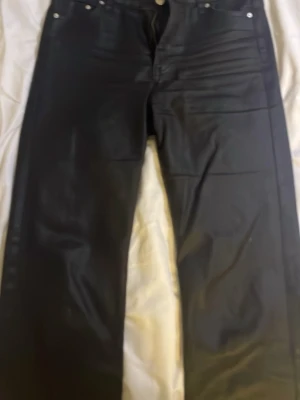 Svarta skinn jeans med rå kant och dragkedja - Säljer ett par svarta skinn jeans med rå, fransig kant längst ner och klassisk femficksdesign. Byxorna har dragkedja och knapp framtill samt bälteshällor. Materialet är jeans och passformen är rak med normal midja.