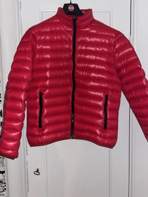 Röd pufferjacka från Peak Performance - Säljer en röd pufferjacka från Peak Performance i storlek L. Jackan har en glansig finish, svarta detaljer och dragkedjor på fickorna. Den är quiltad och har en stilren svart patch på ärmen. Perfekt för kalla dagar när du vill vara både varm och snygg. Sitter snarare som M. Nypris runt 3000. 