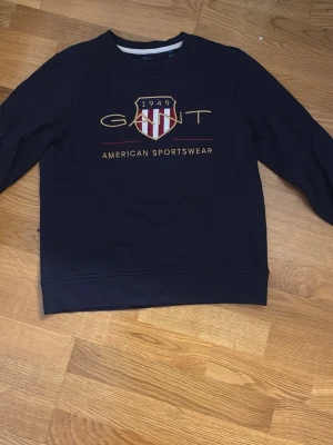 Mörkblå sweatshirt från GANT - Säljer en mörkblå sweatshirt från GANT med stort tryck och broderad logga på bröstet. Tröjan har lång ärm och klassisk rund hals. Perfekt för dig som gillar sportig och stilren design med amerikansk touch.