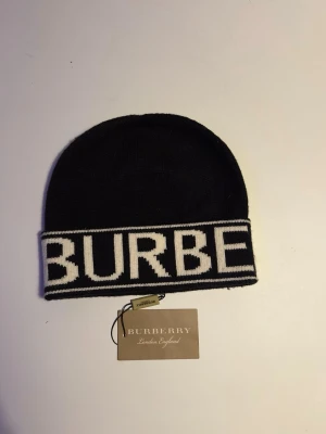 Svart Burberry mössa med logga - Snygg svart mössa från Burberry med bred vit kant och stor stickad logga framtill. Mössan är tillverkad i mjukt material och har en klassisk form som passar perfekt för kalla dagar. Märkesetikett på insidan och originaltagg medföljer.