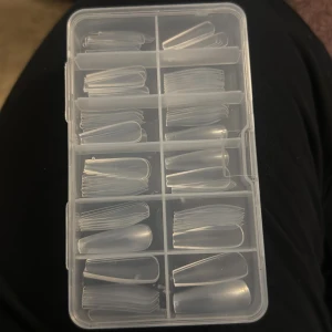 Transparenta lösnaglar i ask - Säljer ett set med transparenta lösnaglar i olika storlekar, organiserade i en praktisk plastask med flera fack. Naglarna är gjorda av plast och passar perfekt för att skapa egna nageldesigner hemma.