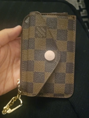 Rutig brun plånbok med kedja - Säljer en snygg liten plånbok  i brunt och beige med klassiskt rutigt mönster. Plånboken har både dragkedja och knapp, samt fack för kort på baksidan och en guldfärgad kedja med karbinhake. Perfekt för kort och småsaker!