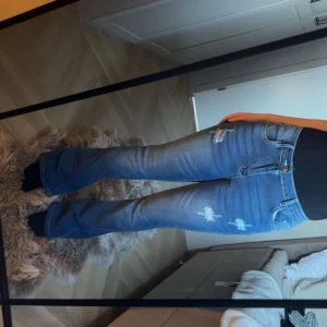 Blå bootcut jeans med slitningar - Säljer ett par blå bootcut jeans med snygga slitningar och ljusare tvätt på låren. Jeansen har klassisk femficksdesign, hög midja och är tillverkade i jeansmaterial med lite stretch. Strlk s-m