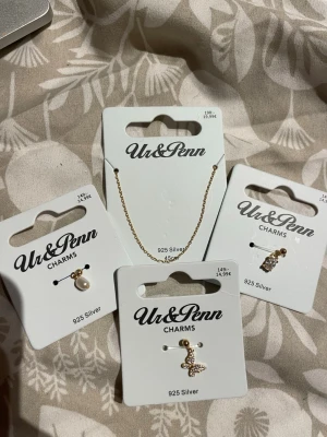 Guldfärgad charmväst från Ur&Penn - Charmig guldfärgad väst med smal kedja och tre olika hängande charms: en vit pärla, en glittrig fjäril och en klar sten. Alla detaljer är i 925 silver med guldfinish, vilket ger en lyxig och modern känsla. Perfekt för att addera lite extra stil till din outfit.