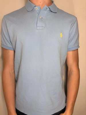 Ljusblå pikétröja från Ralph Lauren - Säljer en ljusblå pikétröja från Ralph Lauren med klassisk krage och två knappar framtill. Tröjan har korta ärmar och den ikoniska gula logon broderad på bröstet. Materialet är mjuk bomull som känns skönt mot huden. Perfekt för dig som gillar stilrena och tidlösa plagg.