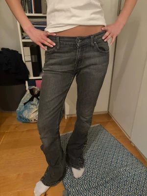 Grå bootcut jeans - Säljer ett par gråa  jeans med låg midja och bootcut-snitt. Jeansen har klassisk femficksdesign, knapp och dragkedja framtill samt slitna detaljer vid bensluten. Perfekt för dig som gillar en avslappnad och cool stil.
