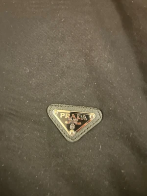 Svart sweatshirt från Prada - Säljer en svart långärmad sweatshirt från Prada med den klassiska metall-loggan på bröstet. Tröjan har rund hals och är gjord i mjukt bomullsmaterial. Perfekt för dig som gillar stilrena designer och exklusiva märken. Storlek s med en skön passform