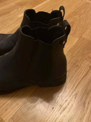Svarta chelsea boots i skinn - Säljer ett par svarta chelsea boots med rund tå och platt sula. Skorna är tillverkade i skinn och har klassiska dragflikar baktill för enkel påtagning. Perfekt för dig som gillar stilrena och tidlösa boots.