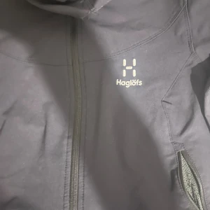 Svart vindjacka från Haglöfs - Svart vindjacka från Haglöfs med huva och hel dragkedja framtill. Jackan har två sidofickor med dragkedja och en stilren design med logga på bröstet. Perfekt för dig som gillar att vara ute och vill ha något funktionellt och snyggt.