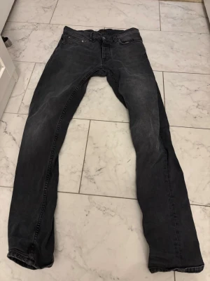 Nudie Jeans, Grim Tim, 31/34 - Säljer ett par svarta jeans från Nudie Jeans Co i storlek 31/34. Jeansen har en klassisk femficksdesign, svarta sömmar och Nudie-logga på bakfickan. Modellen är slim med raka ben och tillverkad i mjukt bomullsmaterial.