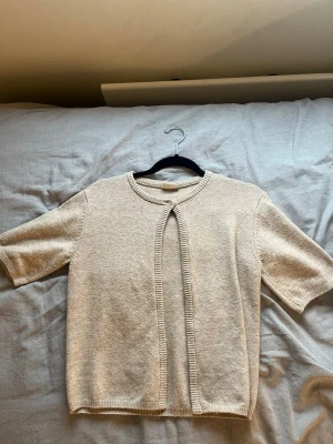 Beige kofta från Kappahl med kort ärm - Beige kofta från Kappahl med korta ärmar och rund halsringning. Koftan har en enkel knapp framtill och ribbade kanter. Perfekt för lager-på-lager och passar till många olika outfits.